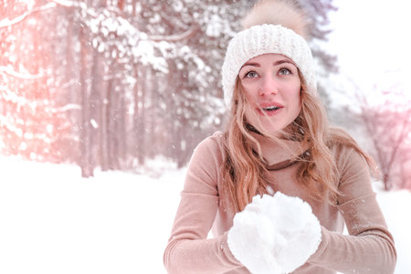 Christmas, holidays and season concept. Young happy woman blowing snow in the winter forest nature. Warm clothing knitted gloves and hat. Winter forest landscape backgroundの写真素材