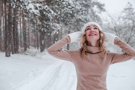 Christmas, holidays and season concept. Young happy woman blowing snow in the winter forest nature. Warm clothing knitted gloves and hat. Winter forest landscape backgroundの写真素材
