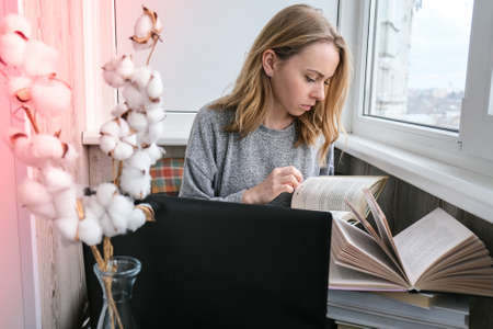 Young woman sitting on balcony reading book, next to laptop. Online education. Stay home concept....