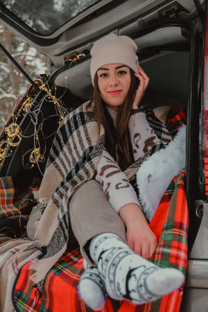Brunette woman wrapped in blanket in trunk car drinking coffee tea from tumbler. Travel in winter. Car decorated with festive Christmas lights. Outdoor picnic. Unity with natureの写真素材