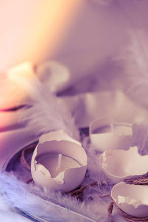 Trendy Easter decorations. Cracked organic eggs on plate. Candle in White shell of the eggs. Bird feather. Copy space. Greeting card. Pastel colors. Easter DIY. Do it yourself.の写真素材