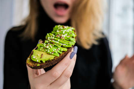 Female hand holding healthy avocado toast. ripe hass avocado, wholegrain bread, sesame flax seeds. Vegan keto diet. Healthy lifestyle. Vegetarian trendy guacamole.の写真素材