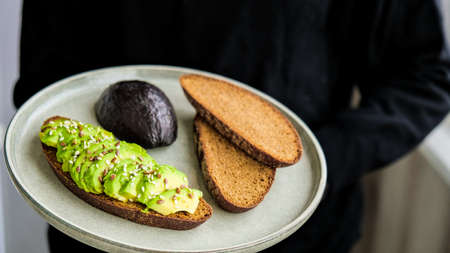 Female hand holding plate with Ingredients for healthy avocado toast. ripe hass avocado, wholegrain...