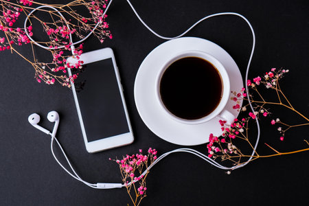 Headphones mobile phone. Black screen. Copy space for text. Top view. Dry pink flowers decoration. Workplace. Black coffeeの写真素材