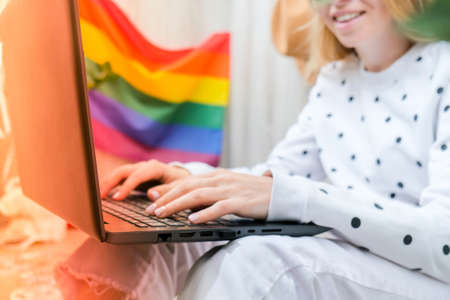 Young millennial hippie woman sitting on balcony using laptop. LGBTQ rainbow flag on background. Online education tuition. Work from home. Modern office. Blonde hair. Stay homeの写真素材