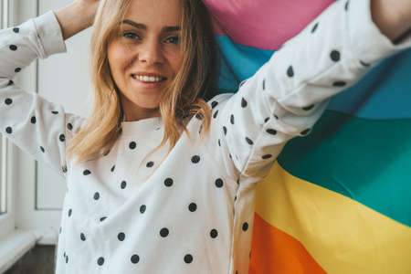 Young blonde millennial hippie woman with rainbow LGBTQ flag at home balcony. Peace and freedom the symbol of LGBT, spending and sharing loving time. Support LGBTQ community. Young lesbian womanの写真素材
