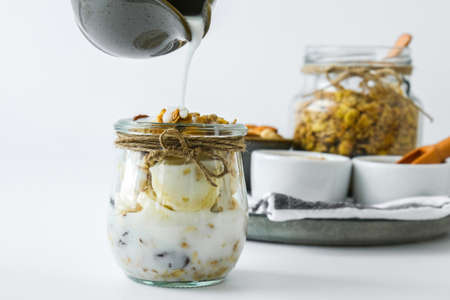 Healthy breakfast. Oatmeal Granola with greek yogurt pouring and nuts banana muesli in jars on light...