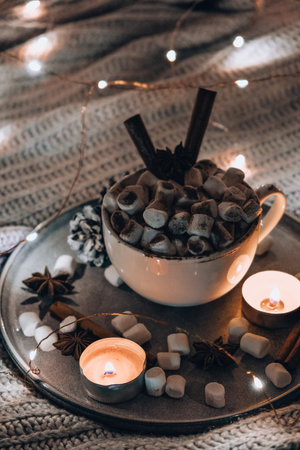 Cup with hot winter cacao and marshmallows cinnamon sticks on bed. Christmas lights. Pine cones...