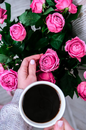 Cup of coffee and pink roses. Holiday concept, bouquet of roses for a birthday, bouquet of flowers in the morning breakfast. Female hands holding cappuccino. Valentines dayの写真素材