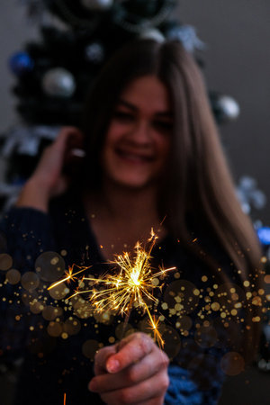 Happy young woman sitting with glass of champagne and bengal light next to the christmas tree. New Year party celebration. Christmas winter holiday.の写真素材