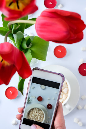Red tulips candles White cup of cacao marshmallows morning breakfast. Female hand taking photo. Blogger. Valentines Womans Mothers dayの写真素材