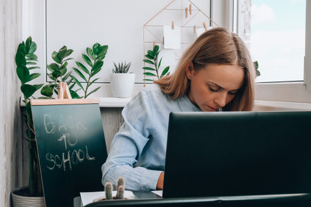 Young female teacher sitting at desk and using laptop at home. e Education Back to school. Student...