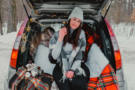 Brunette woman wrapped in blanket in trunk car drinking coffee tea . Travel in winter. Car decorated with festive Christmas lights. Outdoor picnic. Unity with natureの写真素材
