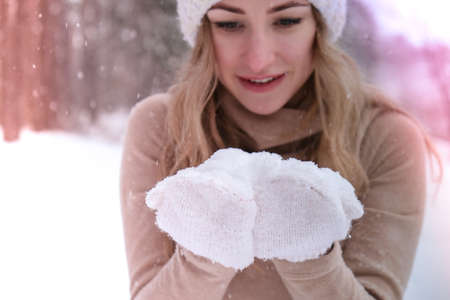 Christmas, holidays and season concept. Young happy woman blowing snow in the winter forest nature. Warm clothing knitted gloves and hat. Winter forest landscape backgroundの写真素材