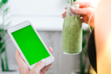 Woman checking smartphone with chroma key green screen drinking vegan smoothie drink detox. Woman doing yoga exercises at home. Online tuition Clean eating, weight loss, healthy dieting food concept. Stay healthy. Wellnessの写真素材