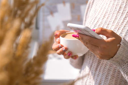 Woman holding Mobile phone. Pampas grass home decoration. Checking social media while Drinking Cappuccino in the breakfast morning at home. Aesthetics Cozy home workplace. Blogger lifestyleの写真素材