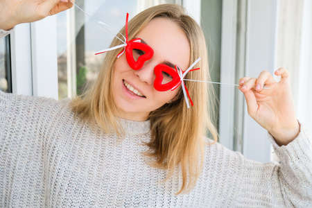 Portrait of beautiful blonde happy woman holding symbol heart. Two red hearts. Girl taking selfie. Alone on Valentines day. Love. Romantic. White sweater, emotionsの写真素材