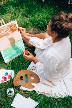 Artist painting on the easel outdoors in the garden. Open air outdoor art workshop. Draw on the canvas with brush and palette sitting on the grass during a picnic in a park. Creativity, art and hobby conceptの写真素材
