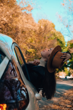 Beautiful young woman carrying hat and looking happy while enjoying road trip. Young woman with long hair in brown hat and sweater looking out from white car holding hat and scarf against autumn forest. Travel vacation road tripの写真素材