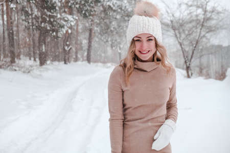 Christmas, holidays and season concept. Young happy woman blowing snow in the winter forest nature. Warm clothing knitted gloves and hat. Winter forest landscape backgroundの写真素材