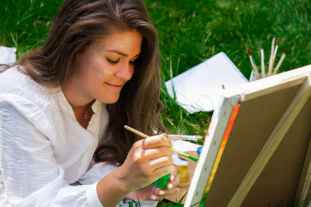 Artist painting on the easel outdoors in the garden. Open air outdoor art workshop. Draw on the canvas with brush and palette sitting on the grass during a picnic in a park. Creativity, art and hobby conceptの写真素材