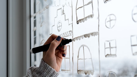 Female hand draws christmas holidays decoration painted on window glass. New Years scenery. city buildings garlands lights. hygge, decoration and christmas concept. festive string at homeの写真素材