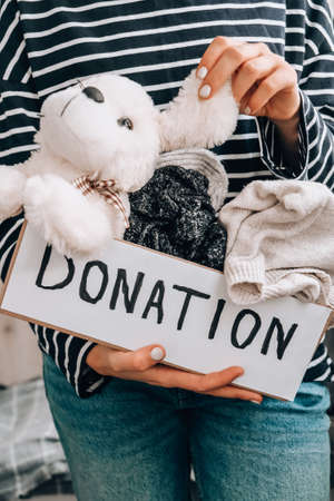 Female volunteer holding donation box with old used toys and clothes indoors. Happy Charity. Unrecognizable woman holding box with clothes in it. close-up. Clothing donation. Winter clothes in a cardboard box. Seasonal clothingの写真素材