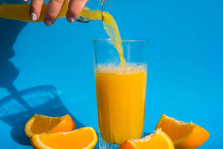 Pouring orange juice stream from jug into glass of squeezing Orange juice with sliced fruits on blue background. Fruit juice, fresh cold drink. Refreshment in summer. Fruits cut in half. Copy spaceの写真素材