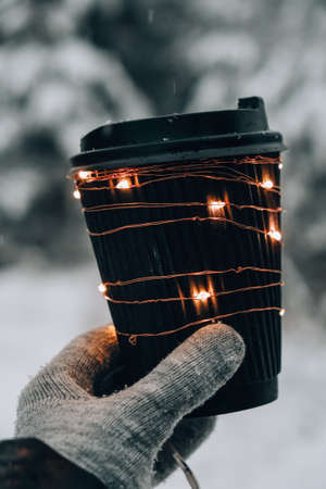 Female hand in winter warm gloves holding black eco paper cup with garland shiny light. Creative trendy zero waste recycle cup. mockup. Hot warming drink to go in winter day. Take away concept leisure concept tea or coffee cupの写真素材