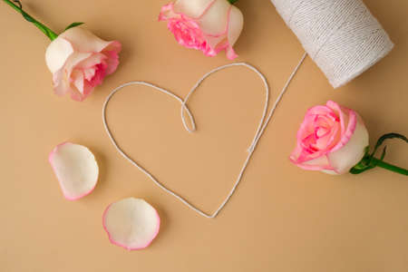 Tender pink roses with spool of white cotton rope in heart shape on beige background. Minimal trendy...