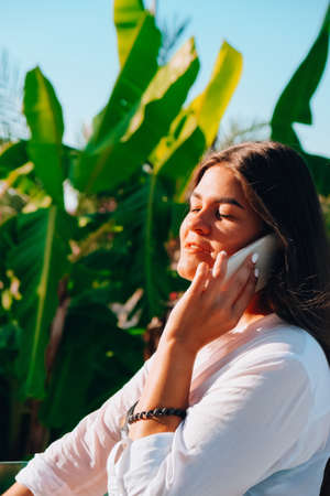 Young beautiful brunette girl talking on her phone on holiday vacations. Tropical palm trees around....