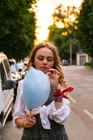 Cute and pretty young girl or student, eats and poses with sugar blue candy cotton on town street, concept happy times. Lifestyleの写真素材