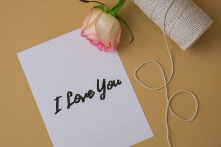 I LOVE YOU text on paper note. Delicate pink roses with spool of white cotton rope on beige background. Minimal trendy composition. Romantic pastel pink rose flowers. Neutral earth tones. Greeting card for Valentines day Modern styleの写真素材