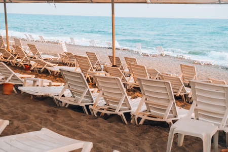 Empty Sun loungers in front of the sea. Sun chairs on sandy beach. Big waves stormy weather. An...