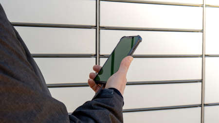Female hand holding mobile phone next to Parcel locker Collecting parcel from shopping locker. Woman...