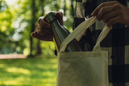 Close-up of Unrecognizable female hand holding Water bottle from eco bag. Reusable steel thermo water bottle in park. Sustainable lifestyle. Plastic free zero waste free living. Go green Environment protection. Woman drinking water in parkの写真素材