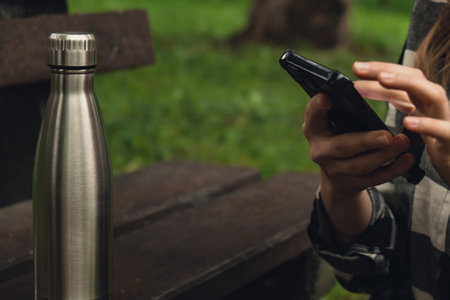 Mobile phone social media. Reusable steel thermo water bottle on wooden bench. Sustainable lifestyle. Plastic free zero waste free living. Go green Woman student work study with phone in parkの写真素材