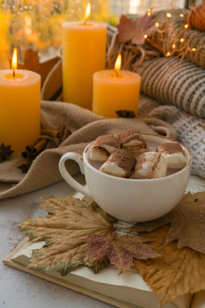 Celebrating autumn holidays at cozy home with cup hot chocolate marshmallows cacao on the windowsill Hygge atmosphere Thanksgiving candle on cozy knitted sweater in warm yellow lights. selective focusの写真素材