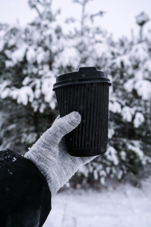 Female hand in winter warm gloves holding black eco paper cup. Winter picnic outdoors. Creative trendy zero waste recycle cup. mockup. Hot warming drink to go in winter day. Take away concept leisure concept tea or coffee cupの写真素材