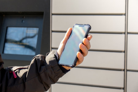 Female hand holding mobile phone next to Parcel locker Collecting parcel from shopping locker. Woman...