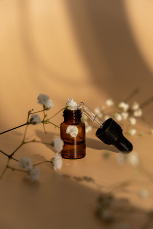 Small bottle of serum on neutral beige background. Trendy shadows. Beauty pipette dropper with Gypsophila or baby's breath white flowers. Glass of cosmetic oil and dried flowers and herbs. Natural organic herbal skin care oil, small flowers. Facial liquid serum with collagen and peptides. Monochrome conceptの写真素材