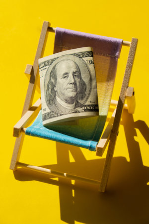 Rolled Up Hundred Dollar Banknote On Miniature Deck Beach Chair. Concept of costs in travel holidays. Save for vacation. copyspace. The concept of how much it costs to restの写真素材