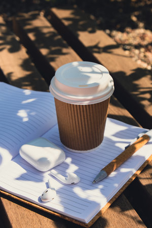 Take away coffee in craft recycling paper cup with paper notebook with wireless headphones. Mockup...