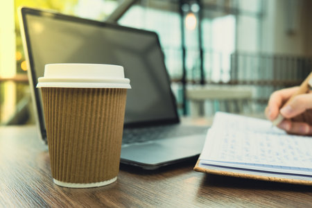 Unrecognizable Young woman study at wooden table in shopping mall food court. Drinking coffee from paper cup. Student making homework Female hands writing on notebook gratitude journal self reflection self discovery in Co-Working Space. Goals, to-do list in the modern office buildingの写真素材