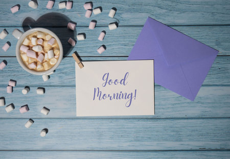 GOOD MORNING text on valentine card inscription positive quote phrase Greeting card with white cup of coffee and marshmallows on wooden blue background. holiday concept. Top view, flat lay minimalistの写真素材