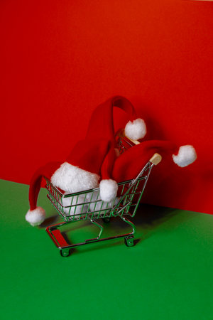 Santa hat in shopping trolley on isometric red green background. Online shopping holiday concept Copy space for your text. Mock up advertisement. Holiday Christmas composition. Minimalist greeting card. Creativeの写真素材