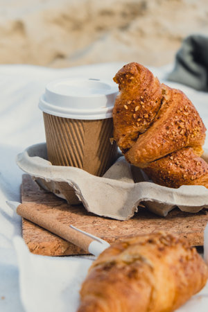 Tasty fresh croissants with coffee in paper cup on sandy beach sea coast background. Breakfast...