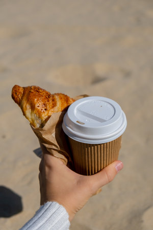Tasty fresh croissants with coffee in paper cup in female hands on beach sea background. Breakfast outdoors wellbeing concept. FPV. Take away foodの写真素材