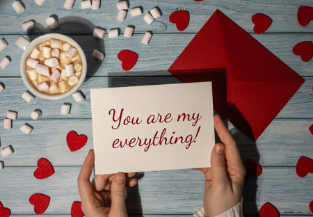 YOU ARE MY EVERYTHING valentine text Female hands holding valentine card red envelope with white cup of coffee and marshmallows on wooden blue background. Romantic Small hearts Valentine day. Holiday morningの写真素材