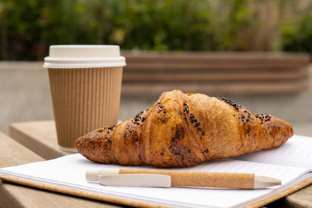 Fresh baked chocolate croissant on paper cup with lid for tea to go. Empty paper notebook....